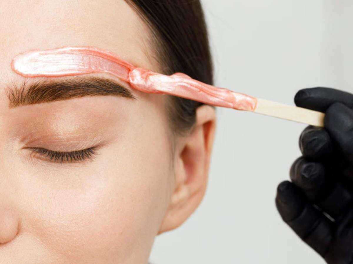 Applying pink wax near eyebrow with a wooden stick held by gloved hand.