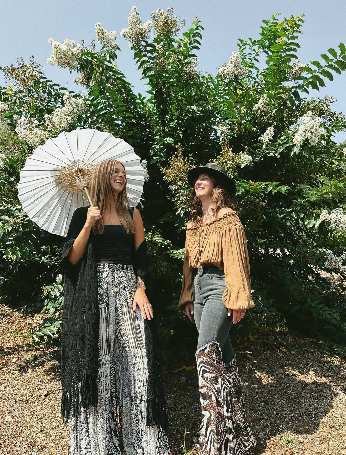 Two women in bohemian attire enjoy a sunny day, one holding a white parasol.